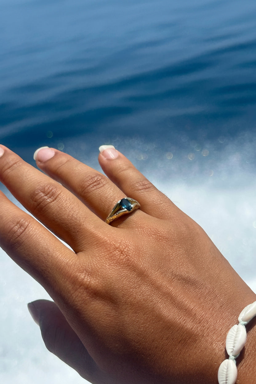 Hand wearing a ring with a blue gemstone against a blurred water background