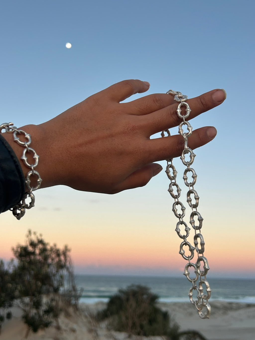 A silver chain bracelet with interlocking ring links worn on a person's wrist and stretched across their fingers, set against a sunset sky and coastal background.