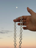 A hand holding up a silver chain with repeating ornate openwork links against a soft blue sky with a visible moon.