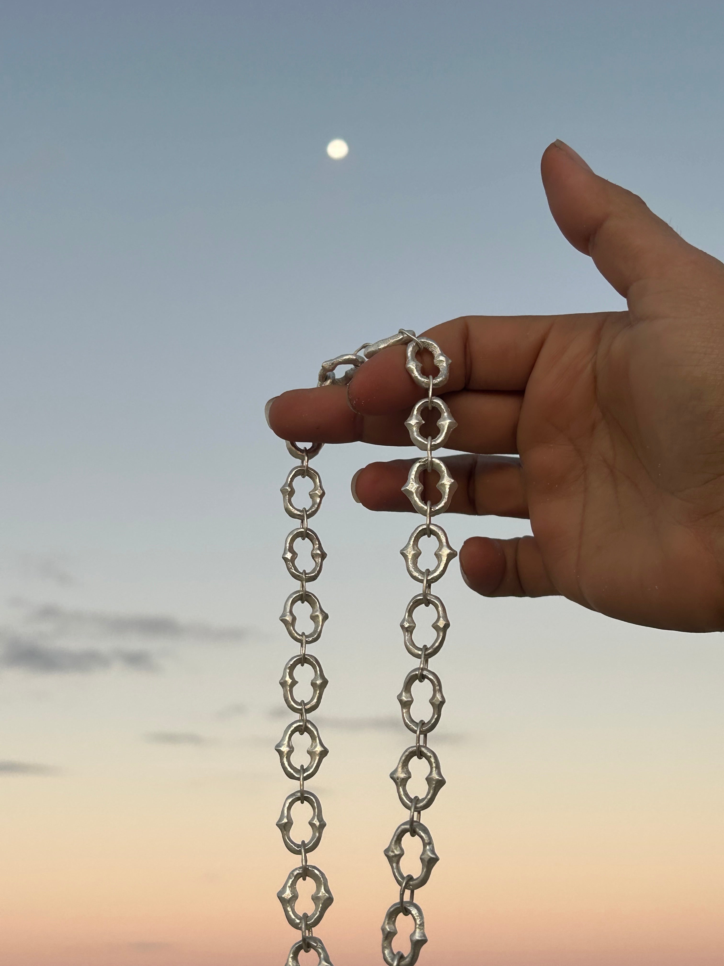 A hand holding up a silver chain with repeating ornate openwork links against a soft blue sky with a visible moon.