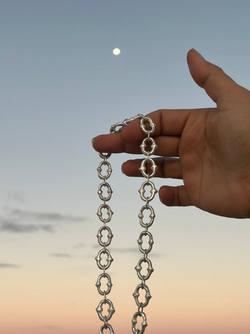A hand holding up a silver chain with repeating ornate openwork links against a soft blue sky with a visible moon.
